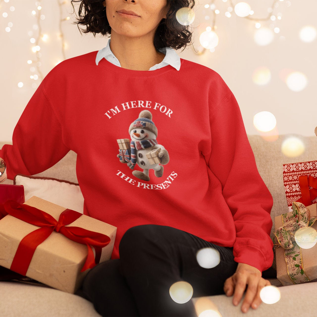 Woman wearing a red sweater with a snowman graphic and text, surrounded by Christmas presents and lights.