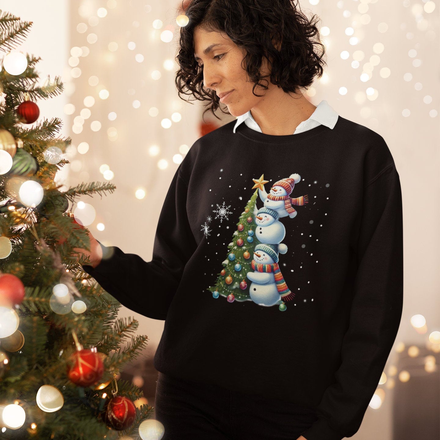 Woman wearing a black Christmas-themed sweater with snowmen and trees, standing next to a decorated Christmas tree.