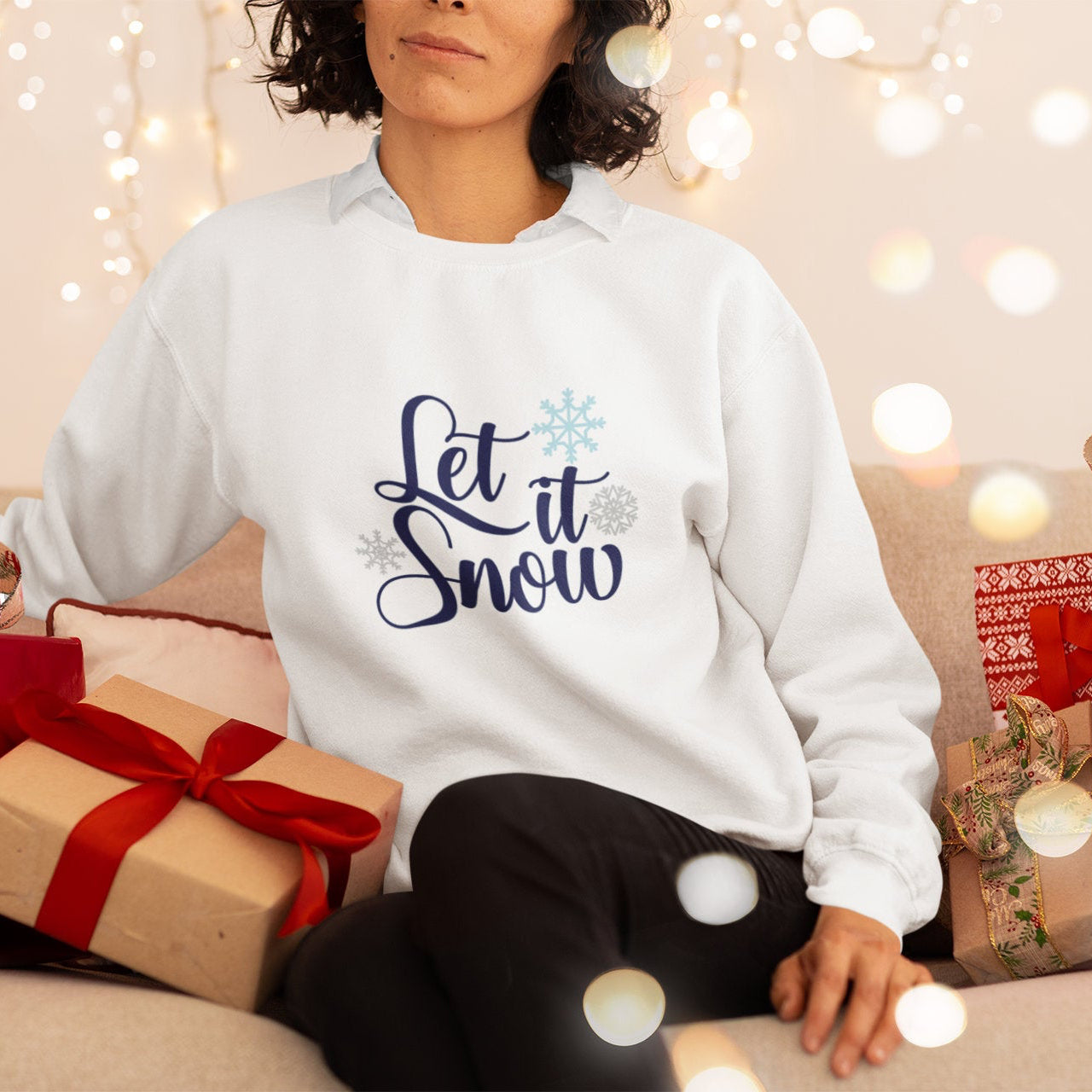 Person wearing a white sweatshirt with 'Let it Snow' text, surrounded by Christmas presents and lights.