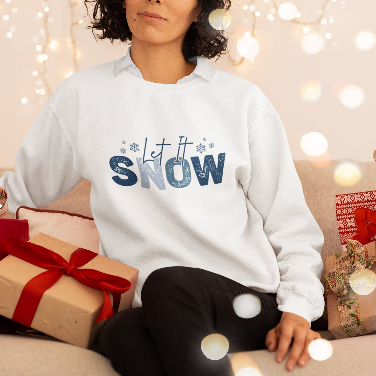 Person wearing a 'Let it Snow' sweater surrounded by Christmas presents and lights.